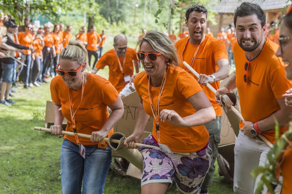 Collègues en tee-shirts orange et lunettes de soleil, hilares, poussant leur char en carton lors d'une course par équipes